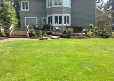 Green backyard with wooden deck, hot tub, and landscaped border beds.